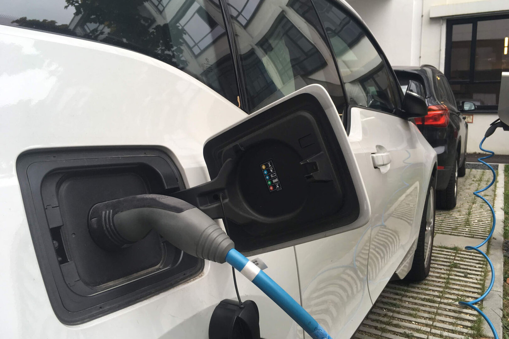 Weißes Elektroauto lädt an Ladesäule angeschlossen, davor im Vordergrund ein Benzinauto