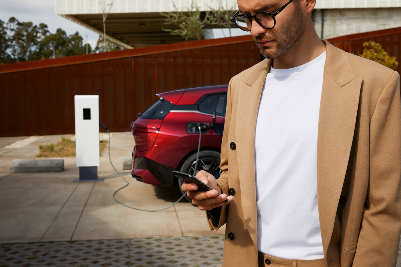 Mann mit Handy in der Hand steht vor privater Ladesäule mit rotem Elektroauto